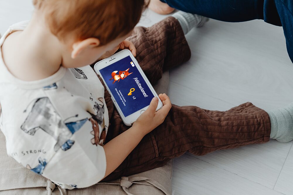 A toddler sits on the floor playing an educational game on a smartphone.